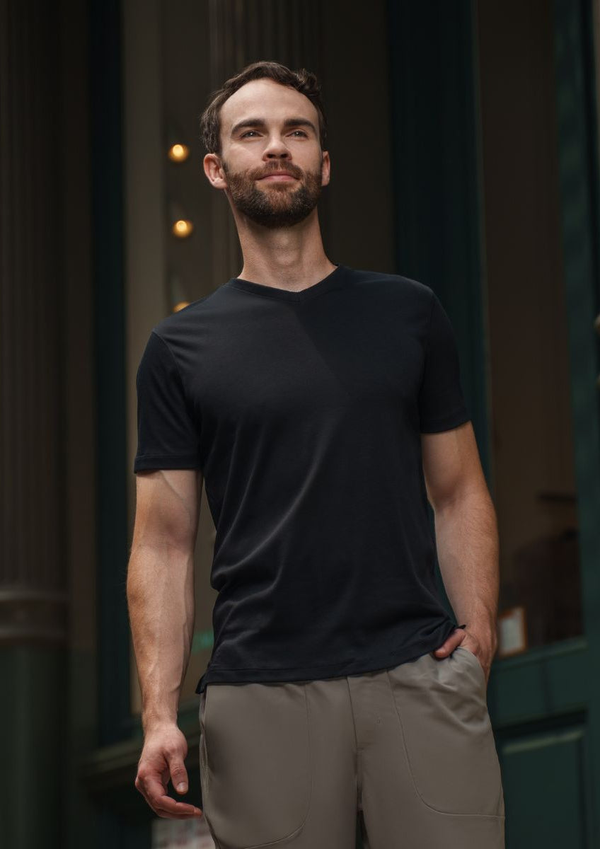 Man wearing a black t-shirt and beige pants standing in front of a dark building entrance.