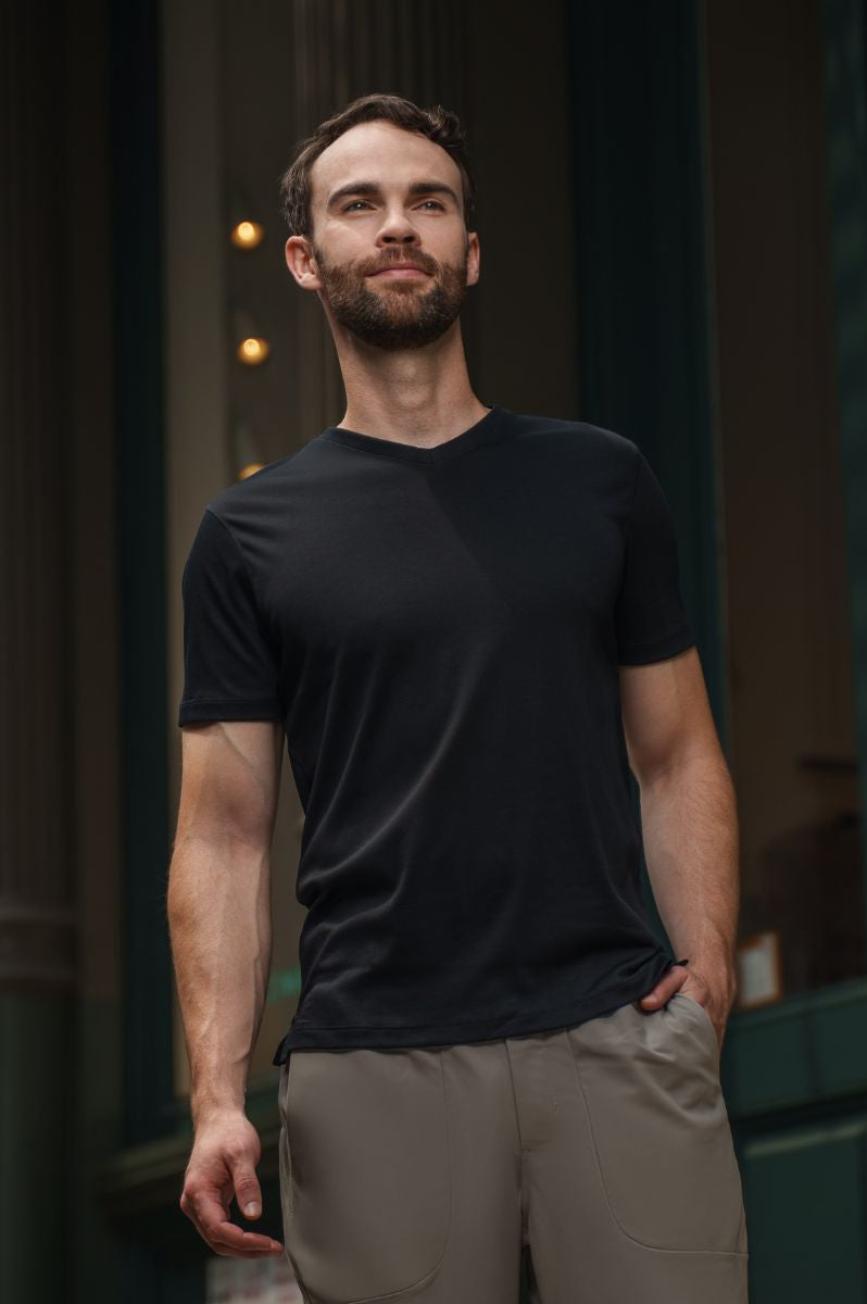 Man wearing a black t-shirt and beige pants standing in front of a dark building entrance.