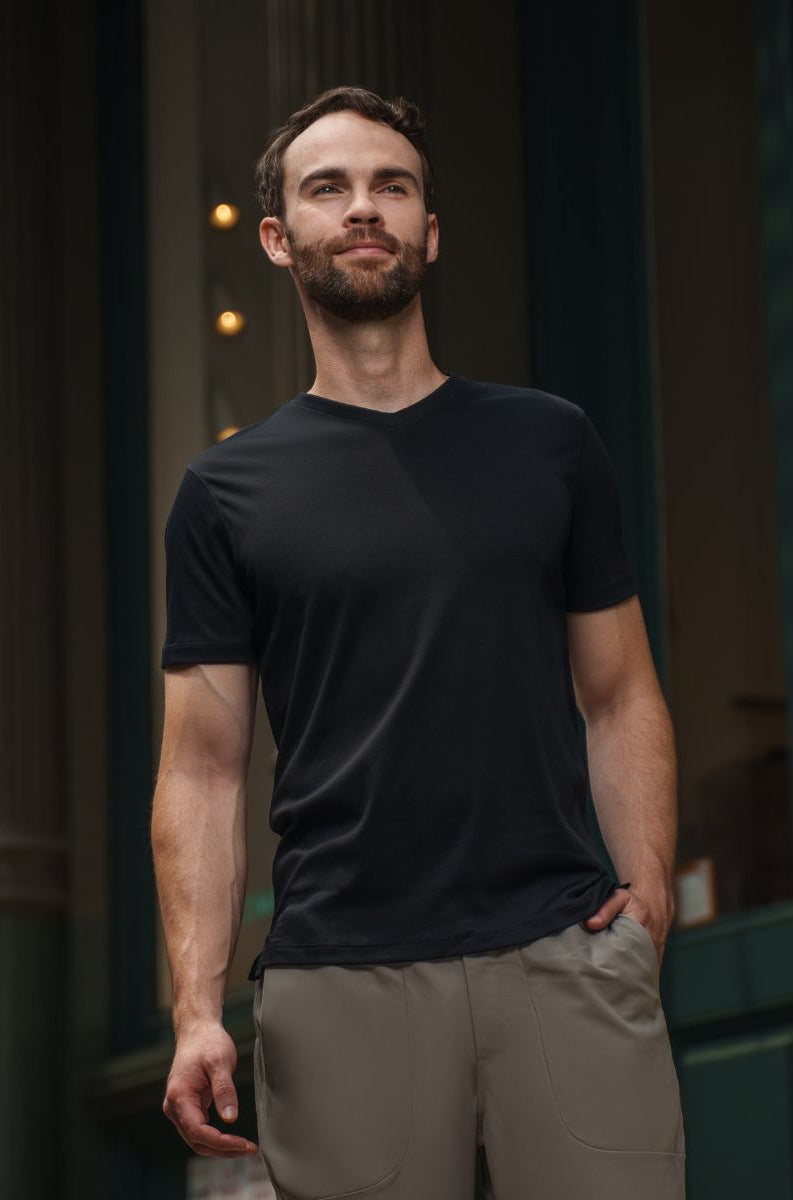Man wearing a black t-shirt and beige pants standing in front of a dark building entrance.