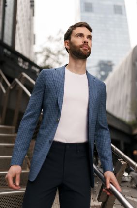 Man in a blue checkered blazer and white shirt standing outdoors with a cityscape background