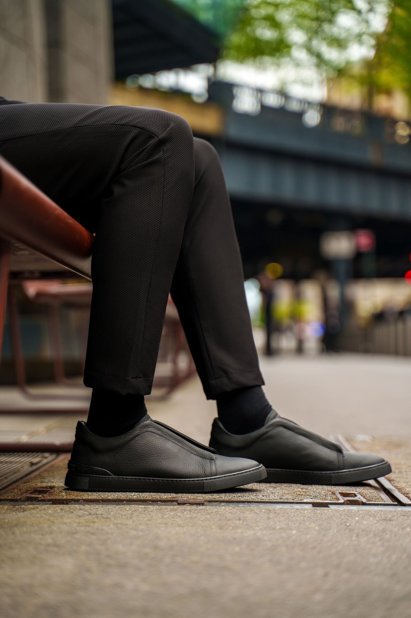 Person wearing black shoes and pants sitting on a bench with a blurred city street background