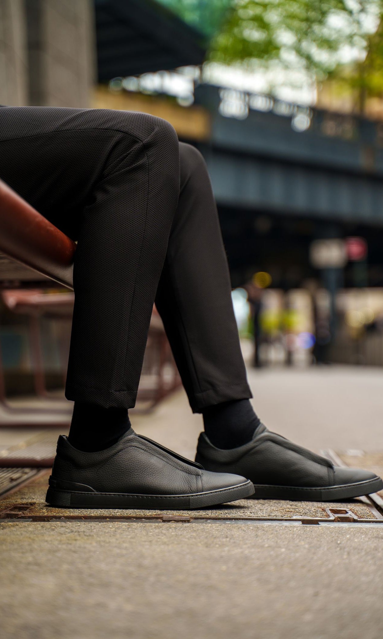 Person wearing black shoes and pants sitting on a bench with a blurred city street background